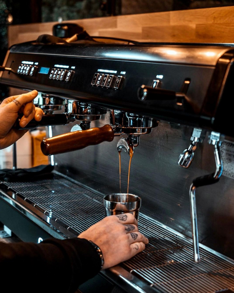 a commercial coffee machine pouring espresso into a shiny cup, with tattooed hands in shot.