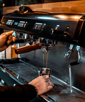 a commercial coffee machine pouring espresso into a shiny cup, with tattooed hands in shot. 