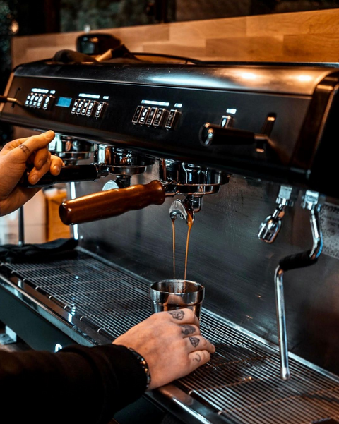 a commercial coffee machine pouring espresso into a shiny cup, with tattooed hands in shot. 